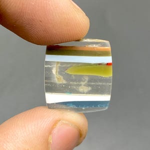 May include: A clear, rectangular cabochon with layered bands of green, brown, white, and blue. The cabochon is translucent and shows the layers of color within.