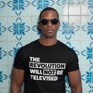 May include: A black t-shirt with white text that reads "THE REVOLUTION WILL NOT BE TELEVISED". The person wearing the shirt is standing in front of a blue and white tiled wall.