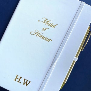 May include: White notebook with a gold elastic closure and a gold pen. The notebook has the text "Maid of Honour" and the initials "H.W." in gold lettering.