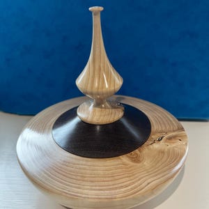 Puede incluir: Una caja de madera con tapa y un remate decorativo. La caja está hecha de madera clara con detalles en madera oscura. El remate es alto y delgado, y está hecho de la misma madera clara que la caja.