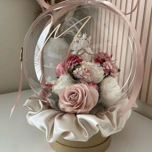 May include: A clear glass globe filled with pink and white flowers, sitting on a beige satin base and a brown box. The globe is tied with a pink ribbon and has the word "Mom" written on it in gold lettering.