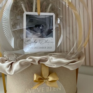 May include: A clear, spherical balloon with a beige satin ribbon and a round, beige box. Inside the balloon is a dried floral arrangement and a photo with the text "Baby Oliver, Due December 2023." A clear tag on the box reads "We've been keeping a little secret!"
