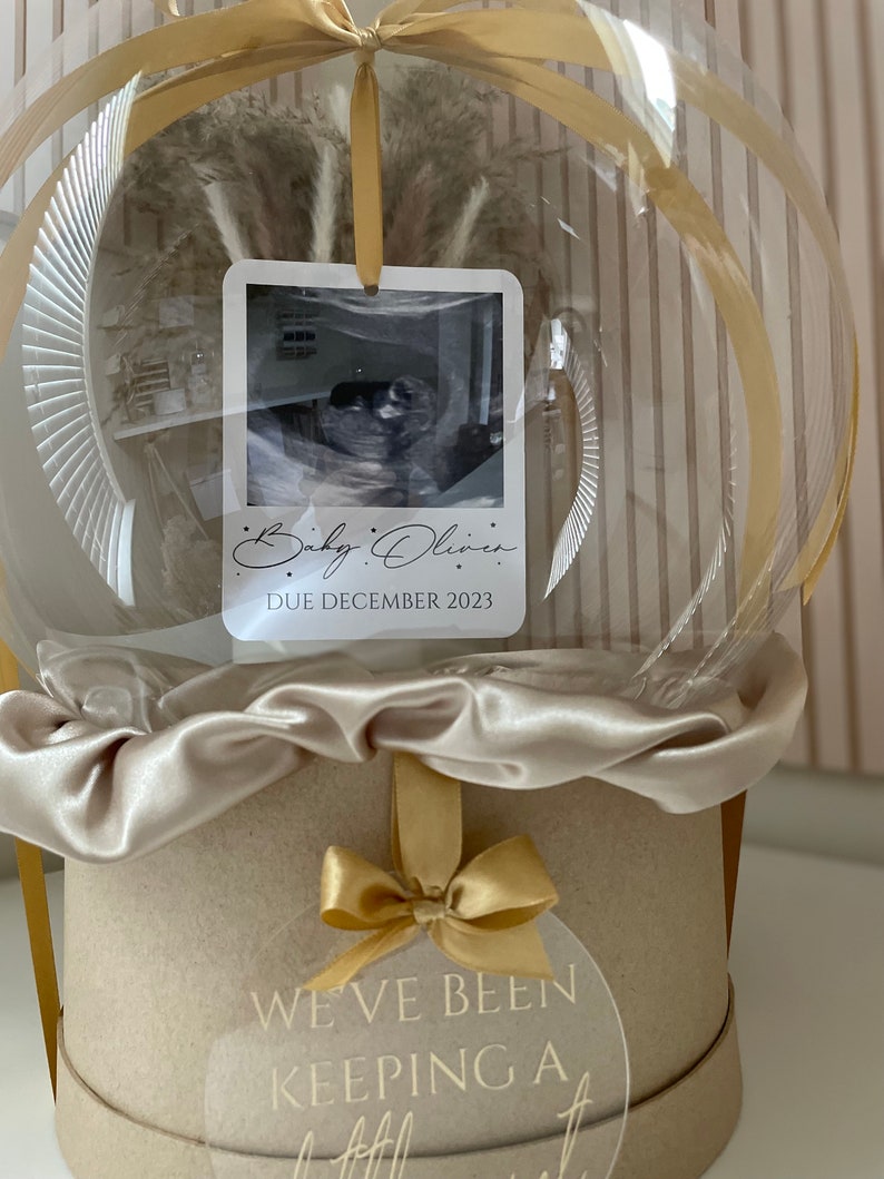 May include: A clear, spherical balloon with a beige satin ribbon and a round, beige box. Inside the balloon is a dried floral arrangement and a photo with the text "Baby Oliver, Due December 2023." A clear tag on the box reads "We've been keeping a little secret!"