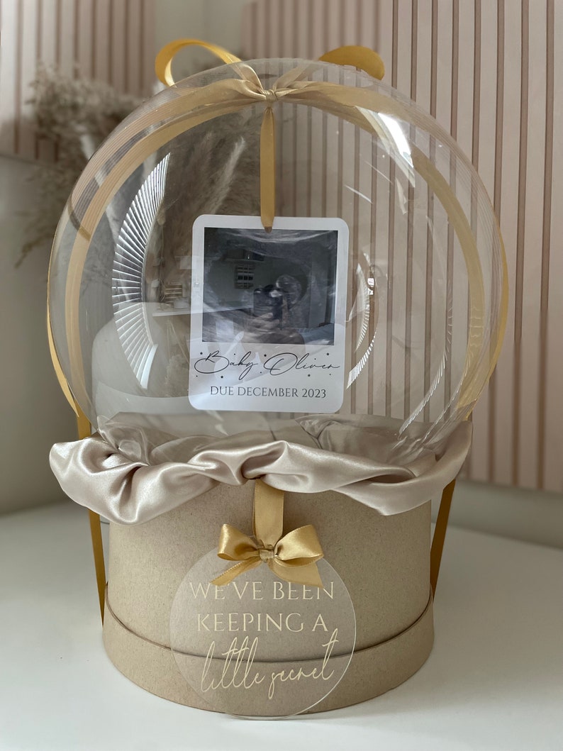 May include: A clear, spherical balloon gift box with a gold ribbon and a round, beige base. Inside, a photo card reads "Baby Olivia, DUE DECEMBER 2023." A circular tag on the base says "WE'VE BEEN KEEPING A little secret."