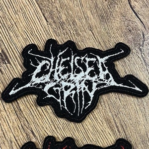 Puede incluir: Dos parches bordados con el nombre de la banda "Chelsea Grin" en estilo black metal. Un parche es blanco sobre negro y el otro es rojo sobre negro.