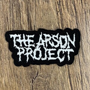 May include: Black and white embroidered patch with the text "THE ARSON PROJECT". The patch has a black border and is set against a wooden background. The text is in a stylized, gothic font.