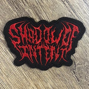 May include: Black embroidered patch featuring the words "Shadow Witch" in red, gothic lettering. The patch has a black border and is on a wooden surface.
