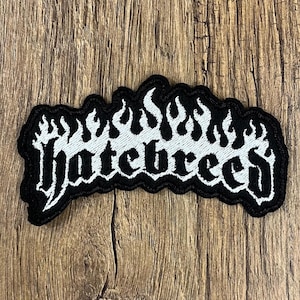 May include: A black and white embroidered patch with the word "Hatebreed" in a stylized font, surrounded by flames. The patch is on a wooden surface.