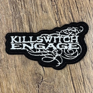 May include: Black and white embroidered patch with the text "KILLSWITCH ENGAGE" in a stylized font. The patch has a black border and is rectangular in shape. The background is a wood grain pattern.
