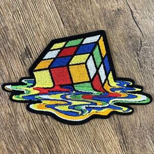 Puede incluir: Un parche bordado que muestra un cubo tridimensional con una cuadrícula de cuadrados rojos, amarillos, azules, verdes y blancos. La base del cubo parece derretirse en un charco irregular y colorido de formas líquidas de colores a juego.