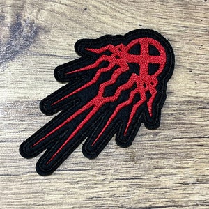 Puede incluir: Parche bordado rojo y negro con un diseño estilizado que se asemeja a un cometa o una estrella fugaz. El diseño está contorneado en negro y relleno de rojo.