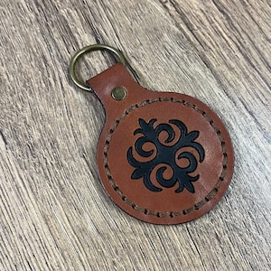 May include: A brown leather keyring with a black, intricate design in the centre. The keyring has a metal ring for attaching keys.