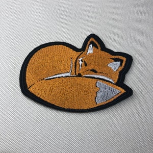 May include: Embroidered iron-on patch of a curled up orange fox with a white tail tip, sleeping on a black background.