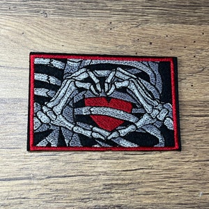 Op de afbeelding: Een geborduurde patch in zwart en wit van skelethandjes die een hart vormen rond een rood hart. De patch heeft een rode rand.