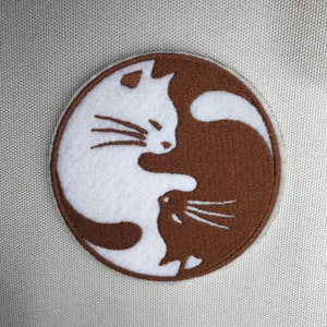 May include: A white and brown embroidered patch featuring two cats in a yin yang design.