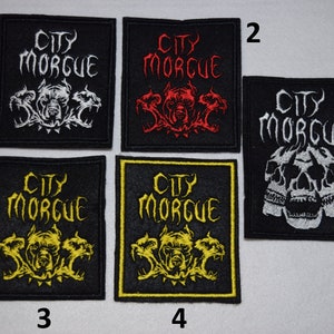Peut inclure: Cinq patchs brodés avec le texte "City Morgue" dans une police gothique noire. Les patchs présentent différents motifs, notamment des chiens à trois têtes, des crânes et un crâne à trois têtes. Les patchs sont numérotés de 1 à 5.