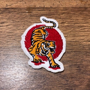 Puede incluir: Un parche de hierro bordado con un tigre con rayas naranjas y negras, de pie sobre un círculo rojo con un contorno blanco.