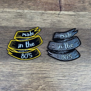 Könnte beinhalten: Zwei gestickte Patches mit dem Text "made in the 80's" auf schwarzem Hintergrund. Ein Patch ist gelb und der andere ist grau.