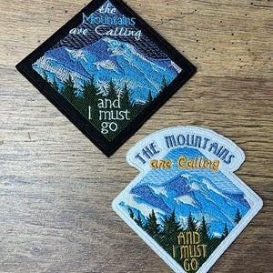 Può includere: Due toppe ricamate con una scena di montagna e il testo "The Mountains are Calling and I must go". La toppa superiore è nera con testo bianco e la toppa inferiore è bianca con testo giallo.