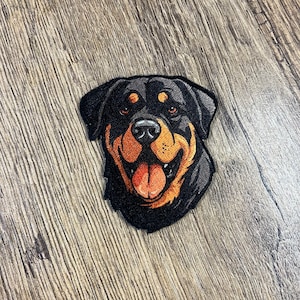 May include: Embroidered patch of a Rottweiler dog's face. The dog has black fur, a brown nose, and a brown tongue sticking out. The dog is smiling.