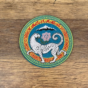 May include: A blue and yellow circular embroidered patch featuring a white snow leopard with black spots, a red and yellow border, and the text "Turan Ayal" in a yellow circle.
