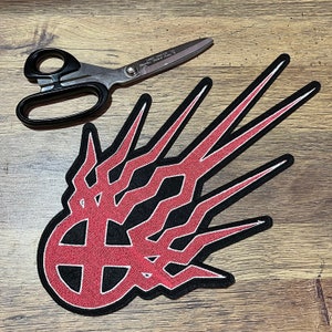May include: Red and black embroidered patch featuring a stylised design of a cross with flames radiating outwards. The patch is made of felt and has a white outline.