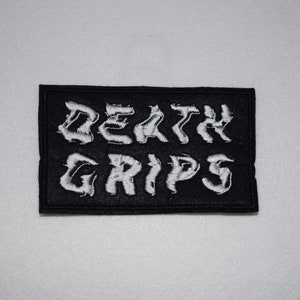 Op de afbeelding: Zwart rechthoekig embleem met de geborduurde tekst "DEATH GRIPS" in witte, gestileerde letters. De patch is waarschijnlijk voor kleding of accessoires.