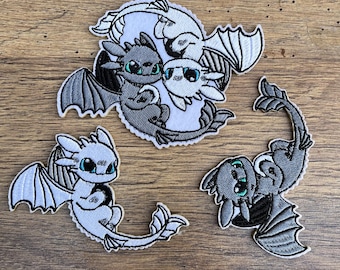 May include: Three embroidered patches featuring cartoon dragons. The first patch shows two dragons, one white and one gray, intertwined in a yin-yang symbol. The second patch shows a white dragon with blue eyes. The third patch shows a gray dragon with blue eyes.