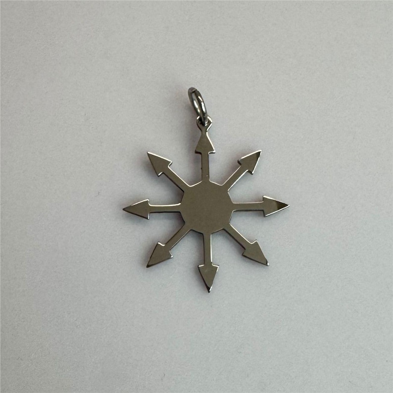 May include: A silver-colored pendant featuring a stylized eight-pointed star design with arrows pointing outwards. The pendant has a small loop at the top for a chain. The pendant is likely made of metal and is displayed against a white background.