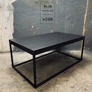 Op de afbeelding: Zwarte salontafel met een rechthoekig houten blad en een zwart metalen frame. De tafel staat op een betonnen vloer.