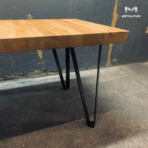 Op de afbeelding: Een houten tafelblad met een natuurlijke houtnerf afwerking rust op zwarte metalen haarspeldpoten. De tafel staat voor een grijze muur.