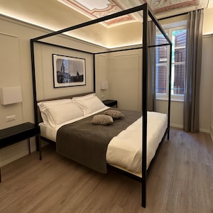 Op de afbeelding: Een moderne slaapkamer met een zwart hemelbed, wit beddengoed en een donkergrijze deken. Twee decoratieve kussens liggen op het bed. De kamer heeft een ingelijste foto, nachtkastjes en een beschilderd plafond met bloemmotieven.