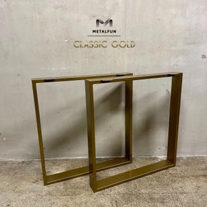 Op de afbeelding: Twee gouden metalen tafelpoten, elk met een vierkant frame, worden getoond tegen een grijze betonnen muur. De poten zijn gemaakt van dik metaal en hebben een gladde, gepolijste afwerking. De tekst "METALFUN CLASSIC GOLD" is zichtbaar bovenaan de afbeelding.