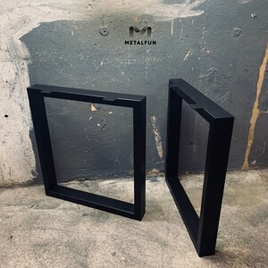 Op de afbeelding: Twee zwarte metalen tafelpoten met een vierkant frame-ontwerp. De poten zijn schuin geplaatst en staan op een betonnen vloer.