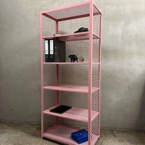 May include: A pink metal shelving unit with five shelves. The unit has a grid-like design on the sides and shelves. Various items are placed on the shelves, including binders, a tablet, and decorative objects. The unit is against a gray wall.