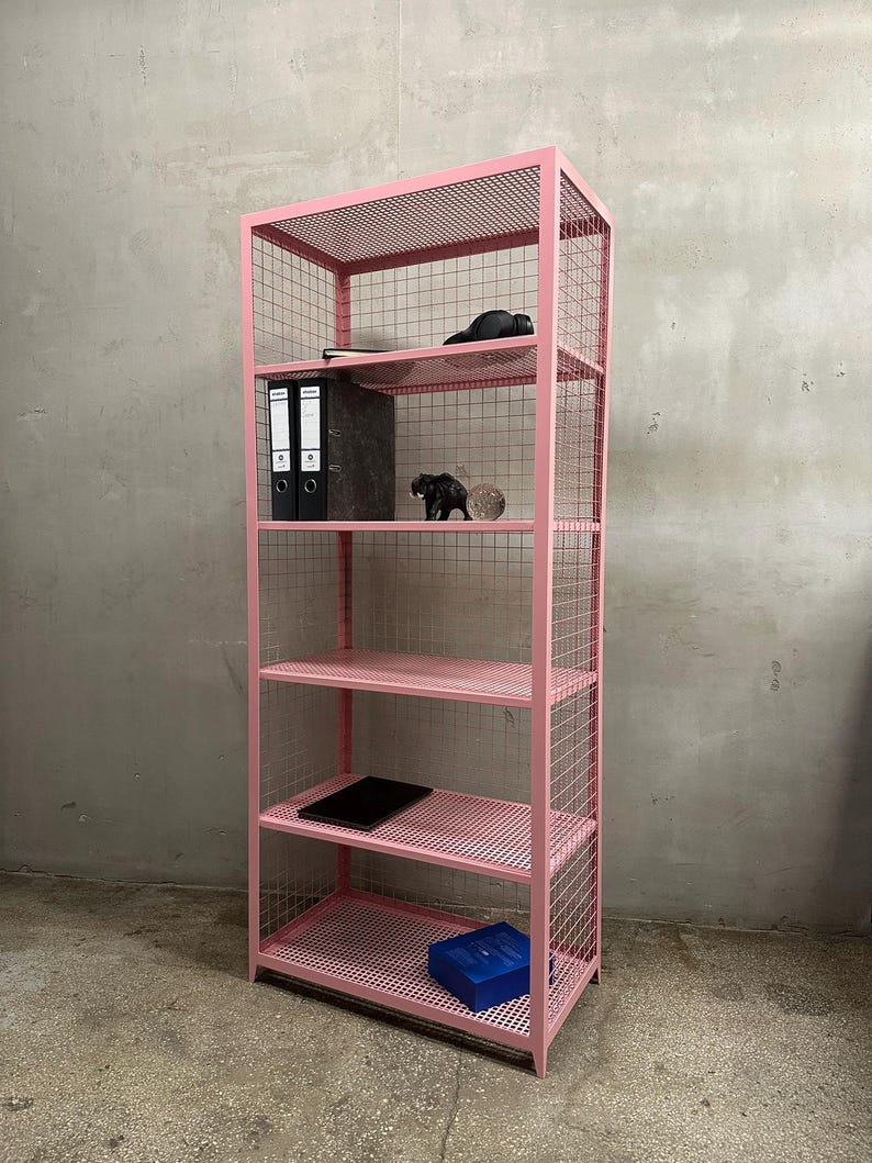 May include: A pink metal shelving unit with five shelves. The unit has a grid-like design on the sides and shelves. Various items are placed on the shelves, including binders, a tablet, and decorative objects. The unit is against a gray wall.