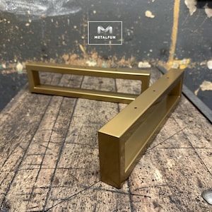 Peut inclure: Deux supports d'étagère en métal doré de forme rectangulaire avec une découpe au centre. Les supports sont posés sur une surface en bois.