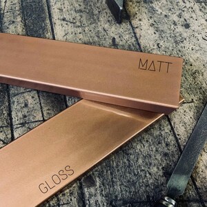 May include: Two rectangular copper metal pieces, one labeled "MATT" and the other "GLOSS". The pieces are a warm copper color and appear to be freshly crafted. A file and a tool are visible in the background, suggesting a workshop setting.