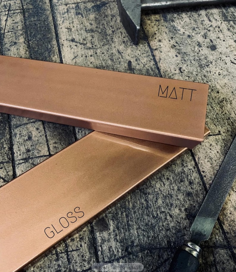 May include: Two rectangular copper metal pieces, one labeled "MATT" and the other "GLOSS". The pieces are a warm copper color and appear to be freshly crafted. A file and a tool are visible in the background, suggesting a workshop setting.