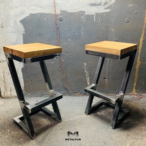 May include: Two industrial-style bar stools. Each stool has a light-colored, rectangular wooden seat atop a metal frame. The metal frame is a dark gray color and has a Z-shaped design. The stools are set against a gray wall. The logo "METAFUN" is in the bottom right corner.