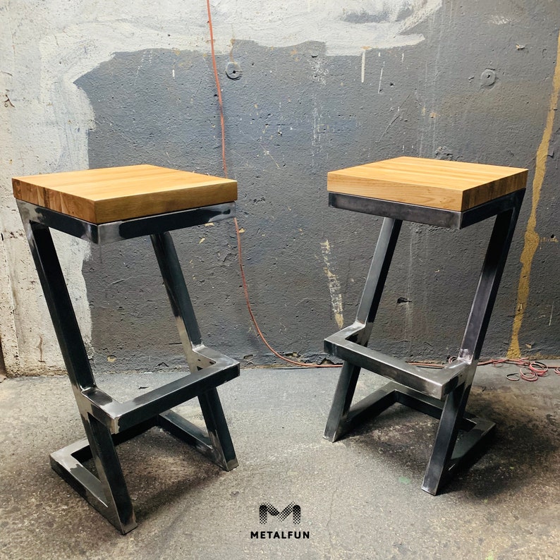 May include: Two industrial-style bar stools. Each stool has a light-colored, rectangular wooden seat atop a metal frame. The metal frame is a dark gray color and has a Z-shaped design. The stools are set against a gray wall. The logo "METAFUN" is in the bottom right corner.