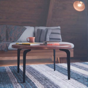 Op de afbeelding: Een houten salontafel met zwarte metalen poten staat op een gestreept tapijt voor een bank. Op de tafel liggen twee boeken en een keramische mok.