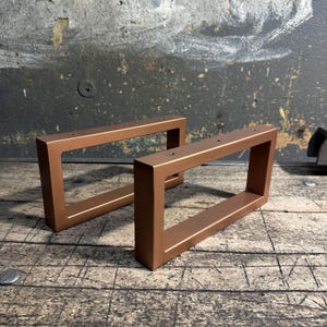 Peut inclure: Deux pieds de table rectangulaires en métal de couleur bronze. Chaque pied a une conception creuse en forme de boîte avec une ouverture rectangulaire. Les pieds sont en métal massif et ont une finition lisse. Ils sont conçus pour le mobilier.
