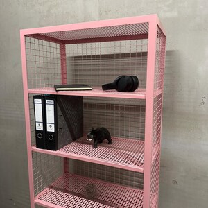 May include: A pink metal wire shelf unit with multiple shelves. The shelves hold a black notebook, headphones, two black binders, a black elephant figurine, and other decorative items. The unit is against a gray wall.
