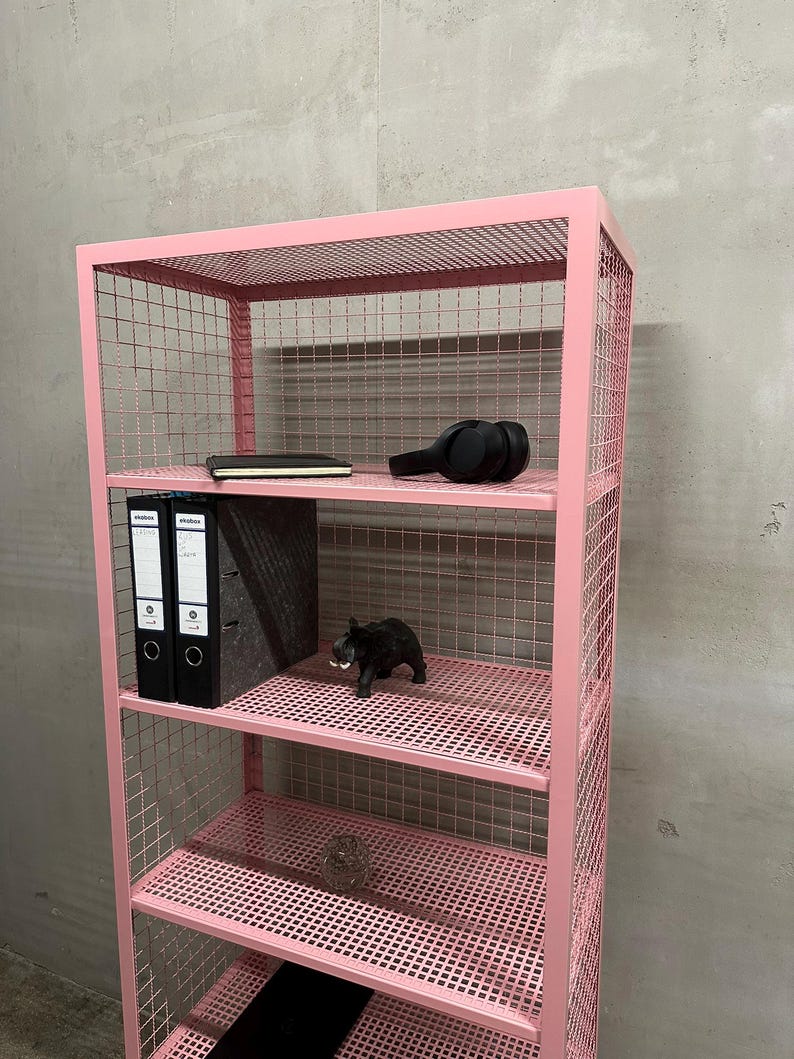 May include: A pink metal wire shelf unit with multiple shelves. The shelves hold a black notebook, headphones, two black binders, a black elephant figurine, and other decorative items. The unit is against a gray wall.
