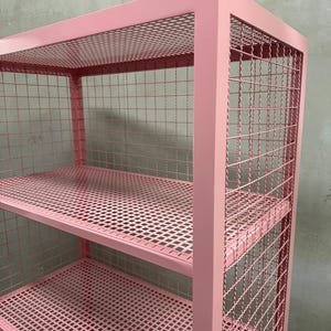 May include: A pink metal shelving unit with three shelves. The shelves and sides are made of a wire mesh. The frame is a solid pink color. The unit is designed for storage and display.
