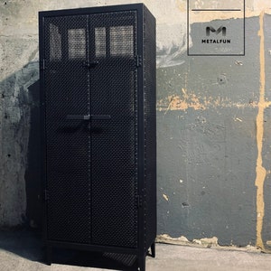 May include: Black metal cabinet with two doors and a wire mesh front. The cabinet has a simple design with a metal handle and hinges. The cabinet is standing on a concrete floor.