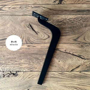 May include: Black metal furniture leg with a curved design. The leg is mounted on a wooden surface.