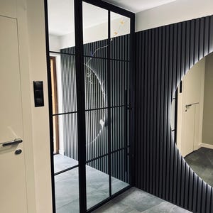 Op de afbeelding: Een hoge glazen deur met een zwart metalen frame en een rasterpatroon, bestaande uit meerdere rechthoekige panelen. De deur staat naast een muur met verticale zwarte latten en een grote ronde spiegel met achtergrondverlichting op een grijze vloer.