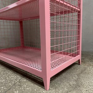 May include: A pink metal storage shelf with three levels. The shelves and sides are made of a wire mesh, and the frame is a solid pink. The legs are angled and the shelf is on a concrete floor.
